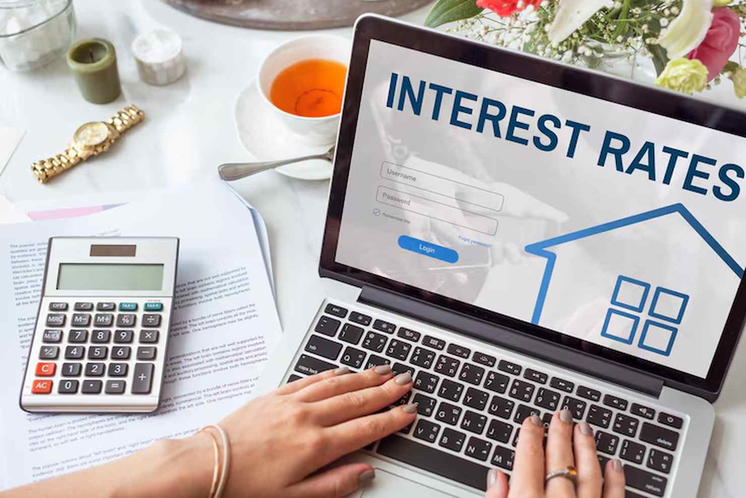 A person typing on a laptop displaying the words "Interest Rates" on the screen, with login fields visible. The workspace includes a calculator, financial documents, a cup of tea, and flowers, suggesting a setting focused on financial planning or loan management.