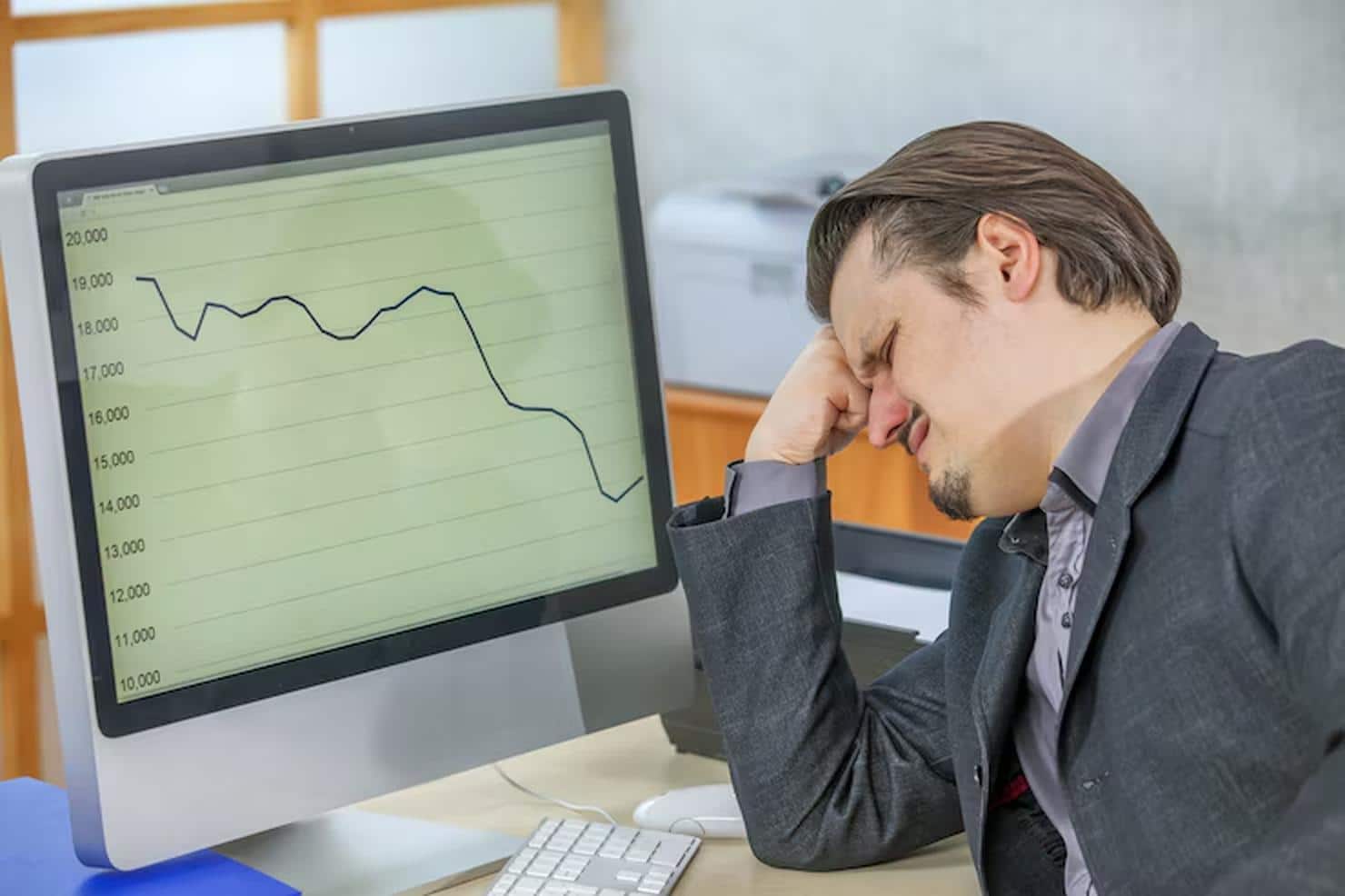 A frustrated businessman sits at his desk with his head in his hand while a line graph on his computer screen shows a sharp downward trend, illustrating the emotional toll and financial stress caused by the inflation impact on markets and personal investments.