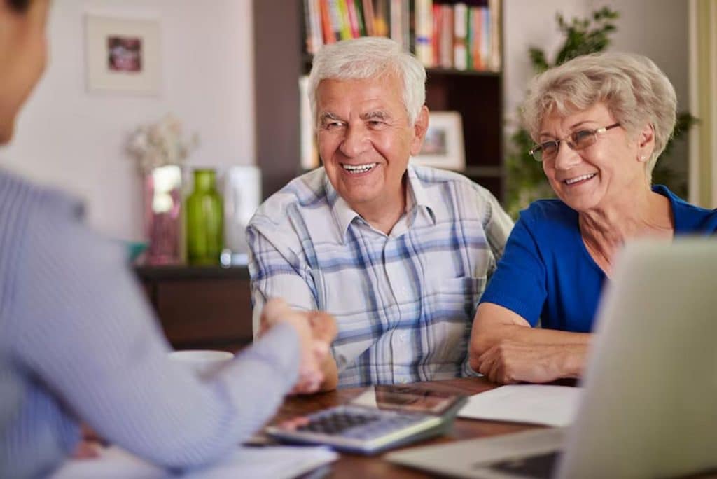 Elderly couple smiling and engaged in conversation with a financial advisor at home, surrounded by documents, a laptop, and a calculator, during a retirement planning session aimed at securing their financial future.