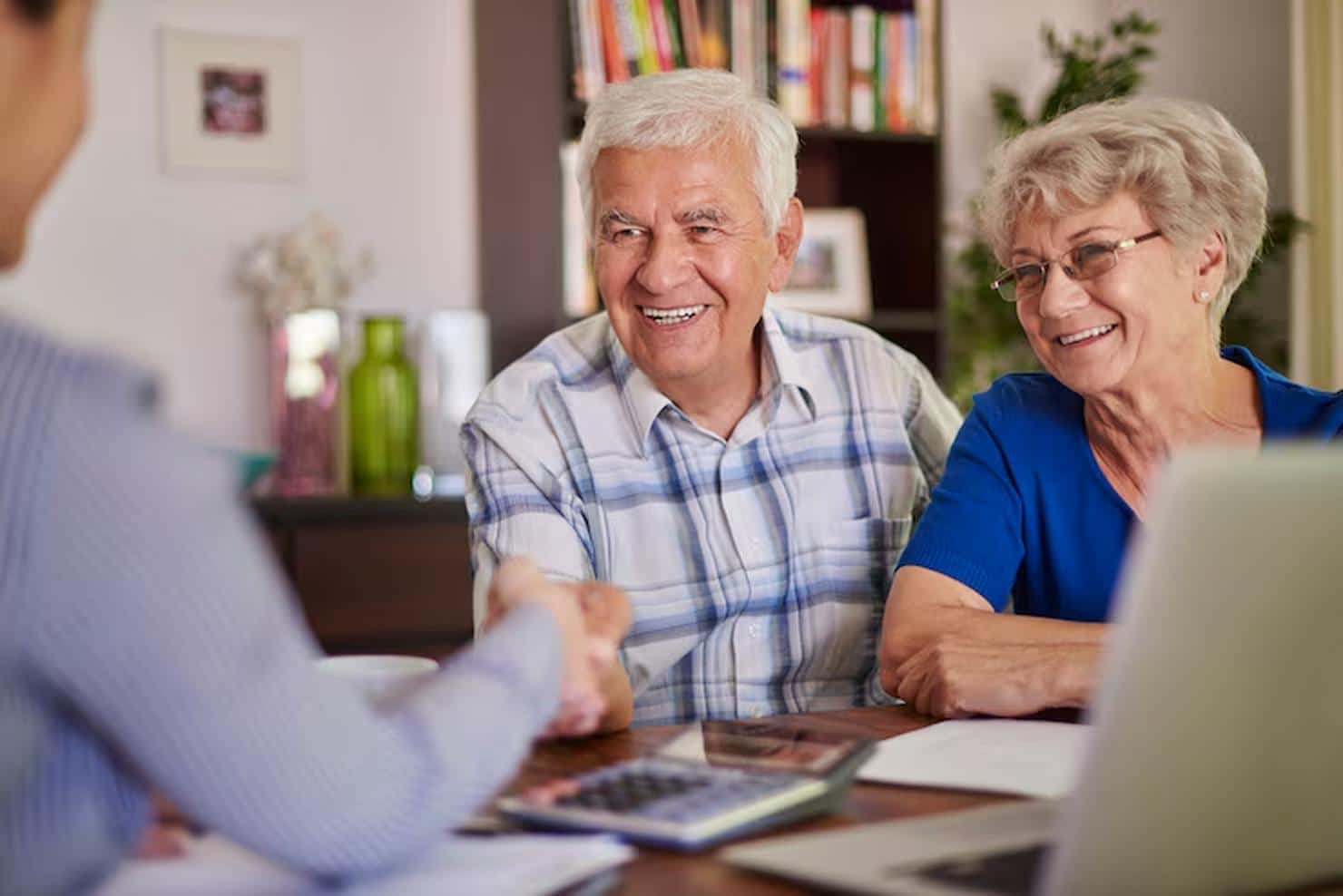 Elderly couple smiling and engaged in conversation with a financial advisor at home, surrounded by documents, a laptop, and a calculator, during a retirement planning session aimed at securing their financial future.