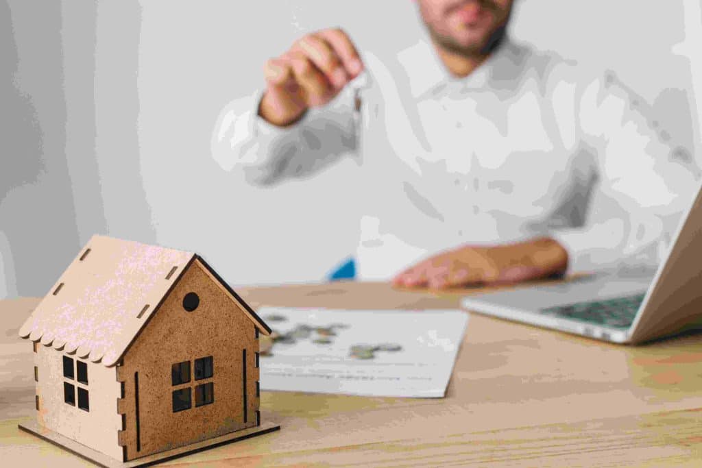 A professional real estate agent handing over house keys at a desk with a miniature wooden house, documents, and a laptop, symbolizing transactions in the estate market.