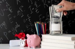 A pink piggy bank, dollar bills in a glass jar, and school supplies on a desk represent financial education for young adults.