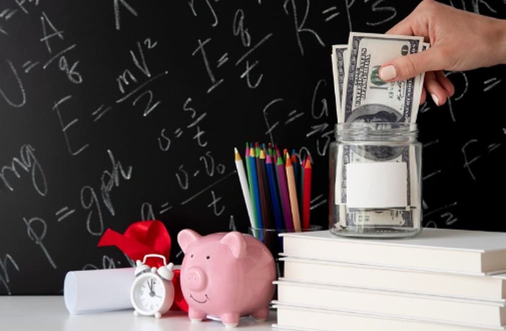 A pink piggy bank, dollar bills in a glass jar, and school supplies on a desk represent financial education for young adults.