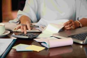 A person sitting at a desk working with a calculator, financial documents, and a laptop, illustrating the process of managing personal debt with careful budgeting and record-keeping.