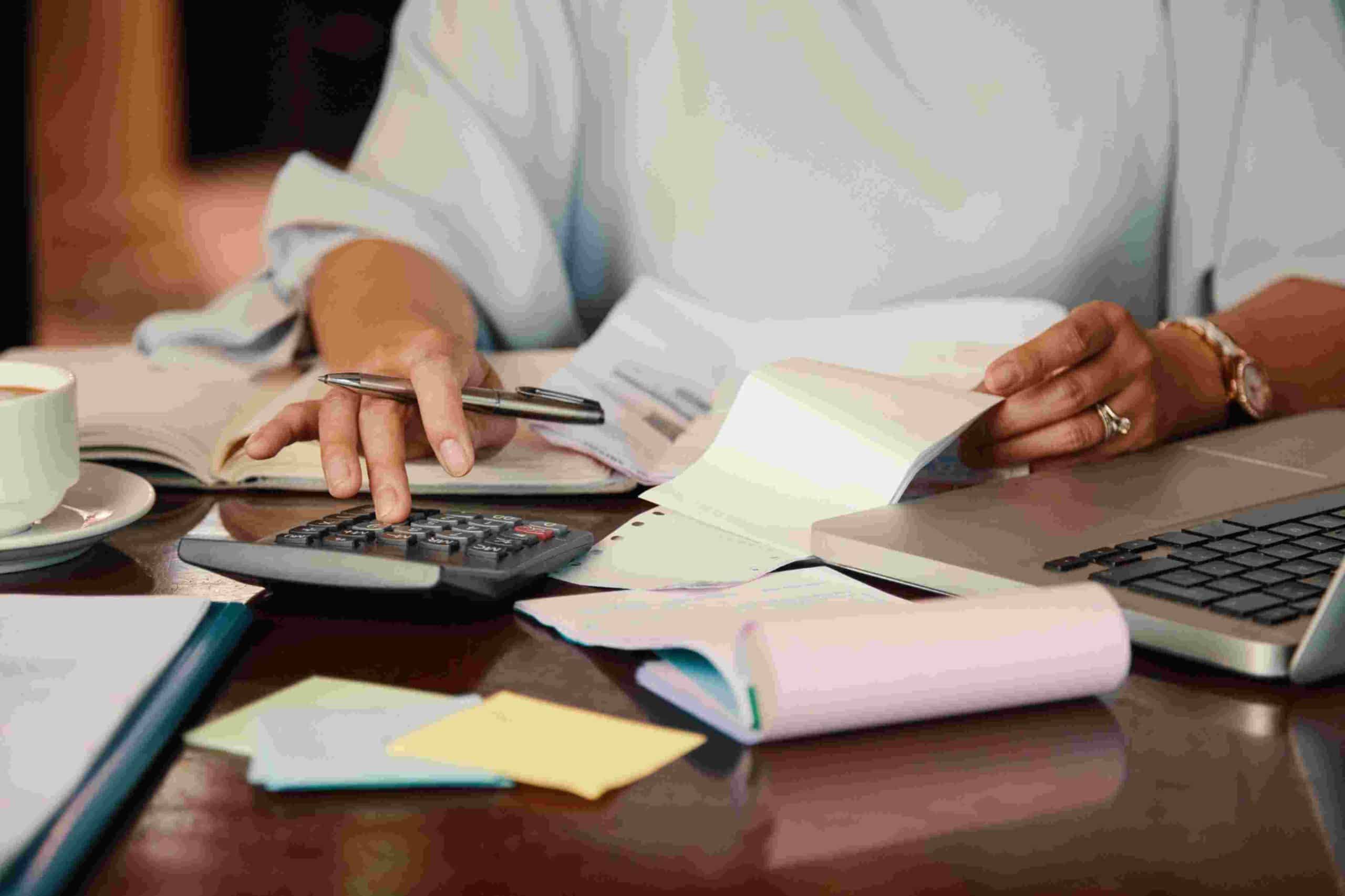 A person sitting at a desk working with a calculator, financial documents, and a laptop, illustrating the process of managing personal debt with careful budgeting and record-keeping.