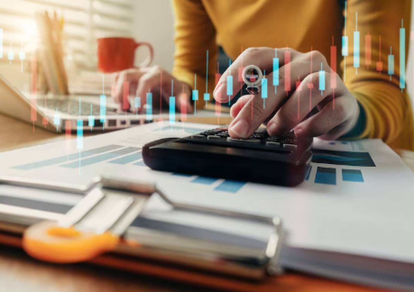 Person using a calculator and laptop to analyze taxes and investments with financial charts and rising bar graphs on a desk.