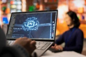 Person working on a laptop displaying a glowing brain graphic with the words “AI” in the center, representing Artificial Intelligence technology development in a modern office setting.