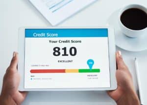Person holding a tablet displaying a credit scoring result of 810 labeled as “Excellent,” with a color scale from bad to excellent, a marker indicating the current score, and a coffee cup on the table nearby.