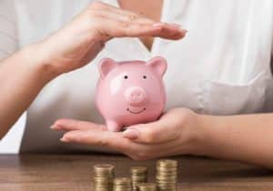 A person protecting a pink piggy bank with their hands, symbolizing savings security and the importance of building emergency funds, with stacks of coins placed on the table in front.