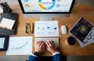 Top view of a professional working on financial planning with charts, graphs, and reports displayed on a computer screen, tablet, and printed documents on a wooden desk.