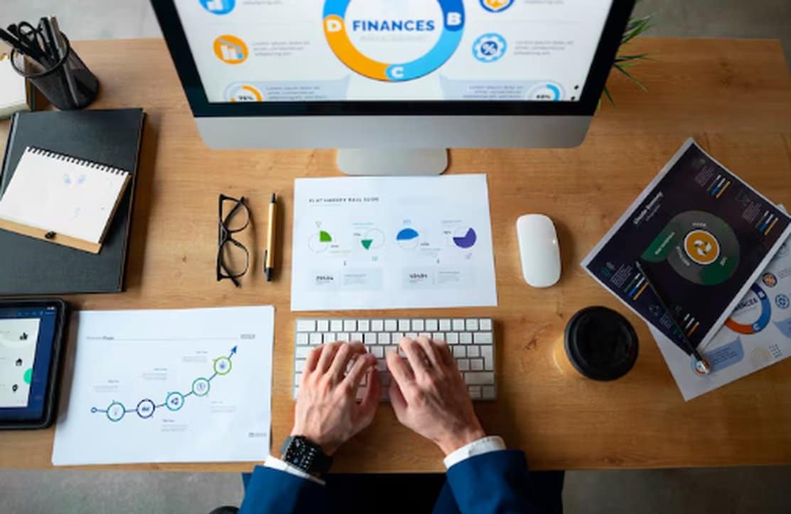 Top view of a professional working on financial planning with charts, graphs, and reports displayed on a computer screen, tablet, and printed documents on a wooden desk.