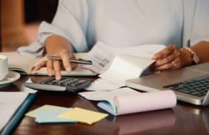 Person calculating investment taxes with a calculator, financial documents, and a laptop on a desk.