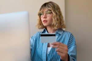 Young woman with glasses shopping online using a credit card in front of her computer, illustrating concerns about invisible bank fees during digital transactions.