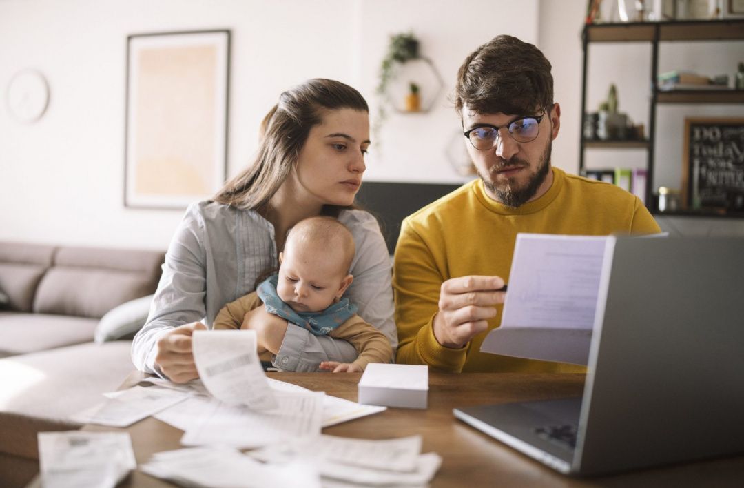 A young couple with their baby sitting at a table, reviewing bills and financial papers while working on their laptop, carefully adjusting their household budgets.