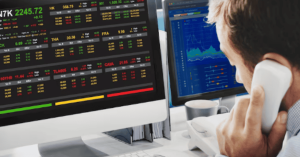 Financial analyst on the phone looking at multiple computer monitors displaying stock prices and market information related to finances.
