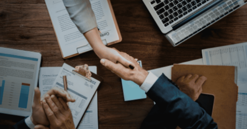 Business professionals shaking hands over a desk with reports, charts, and a laptop displaying business information, symbolizing agreement or partnership.