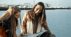 Two women outdoors near the water, looking at documents or maps together, sharing environmental information and collaboration ideas.