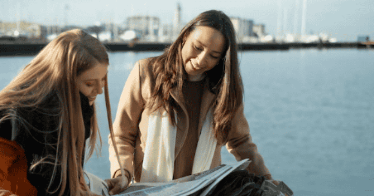 Two women outdoors near the water, looking at documents or maps together, sharing environmental information and collaboration ideas.