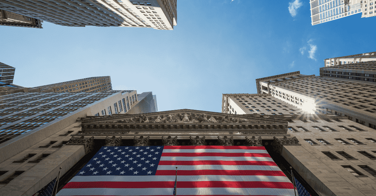 Upward view of Wall Street buildings surrounding a large American flag displayed on a historic financial building, symbolizing information about U.S. markets, economy, and financial institutions.