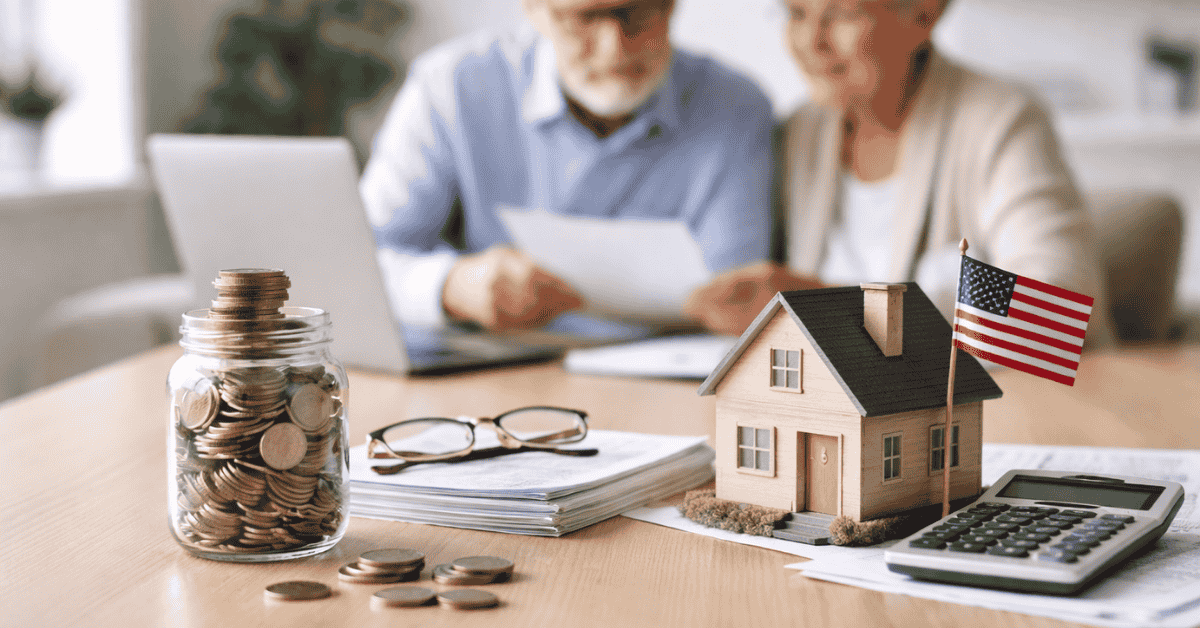 Elderly couple reviewing documents with coins, house model and calculator on table, representing financial information and planning at home.
